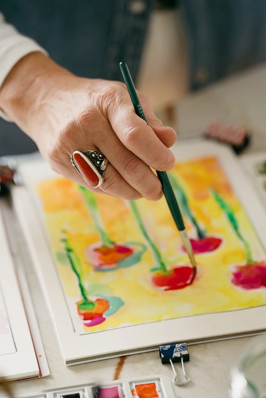 Close-up of a person painting with a thin brush on watercolor paper, which displays colorful abstract patterns, with various art supplies in the background.