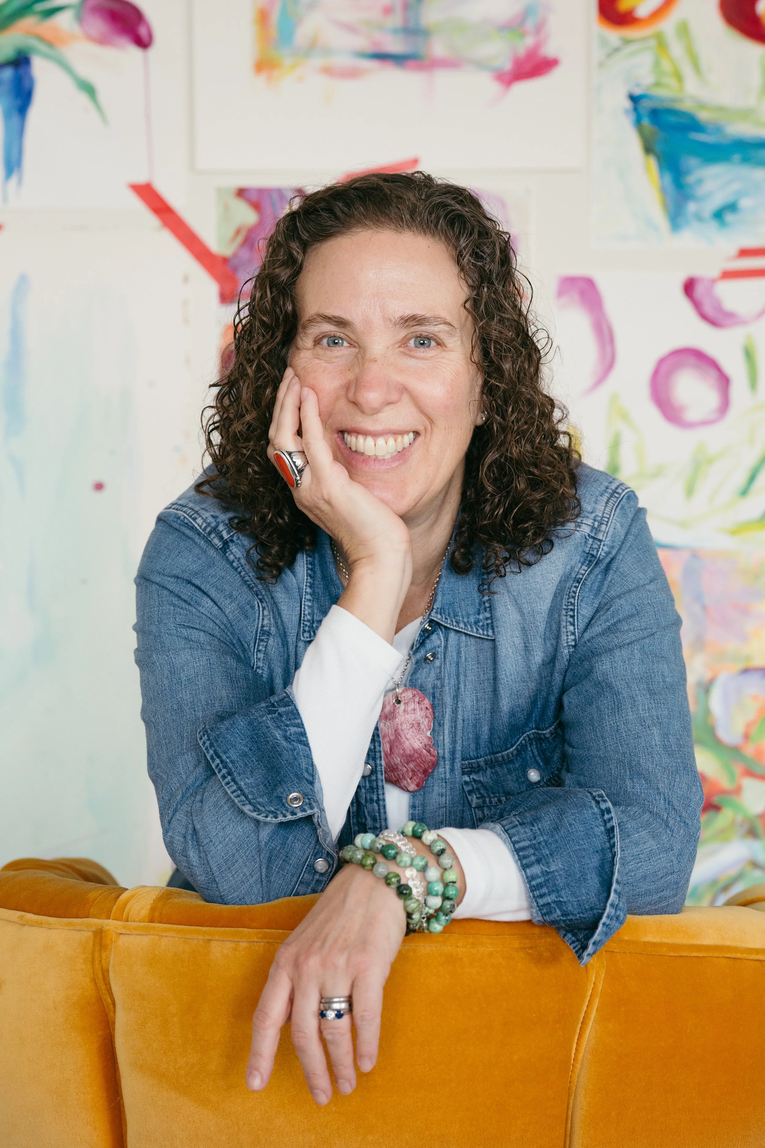 A smiling woman with curly brown hair, wearing a denim jacket, sitting in front of colorful abstract paintings, resting her chin on her hand, featuring jewelry including rings, bracelets, and a large pendant necklace, with an orange sofa in the foreground.
