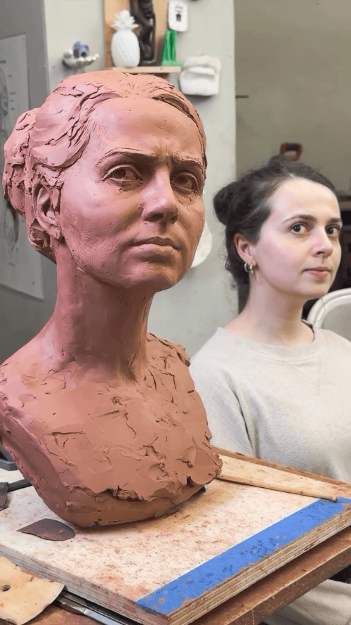 Clay sculpture of a woman's head in progress, with a young woman sitting nearby in an art studio.