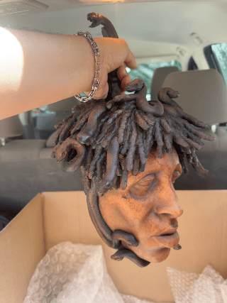 Hand holding a dreadlock-style sculpture of a person's head with detailed facial features inside a box.