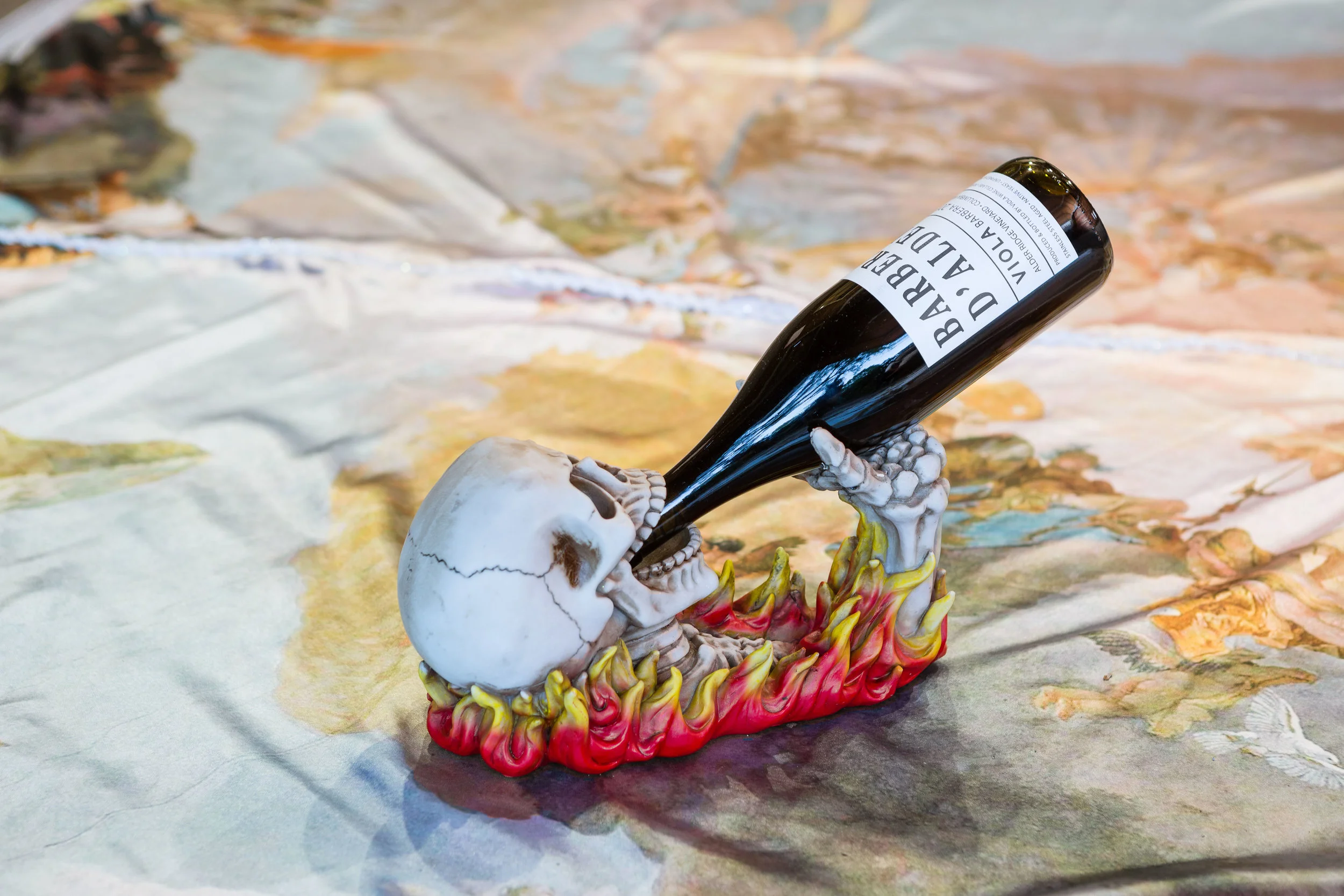 A Soldier, a Magician, and Two Figures Watching a Burning Skull , 2016 (detail view) Oil on linen on three panels, text on lithograph edition, digitally printed satin, sequins, wood, satin cord, flaming skull wine bottle holder, 2014 Barbera d’Alder Viola Wine Cellars Installation view at Cherry | Lucic. Photography by Cristin Norine.