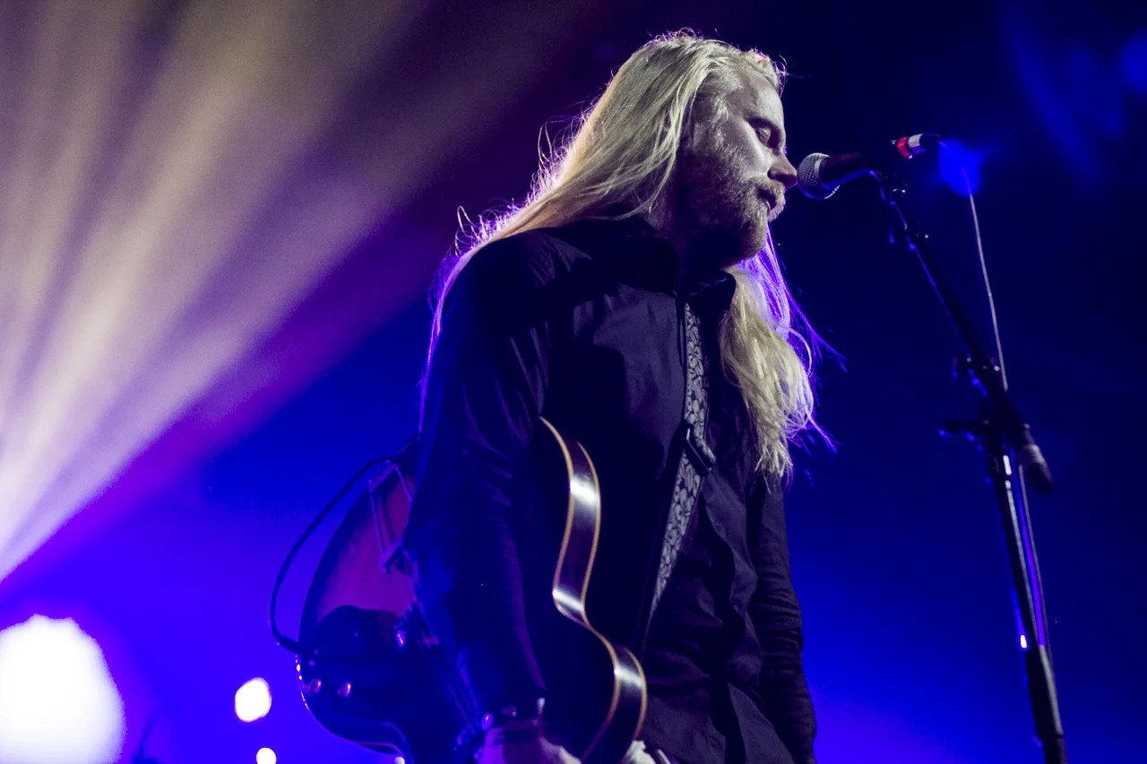 A man with long blonde hair and a beard playing an acoustic guitar on stage with purple and blue lighting, singing into a microphone.