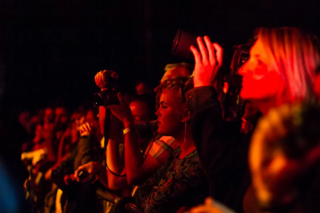 Crowd of people attending a concert or event, with one woman using a camera and others watching and taking photos, illuminated by red lighting.
