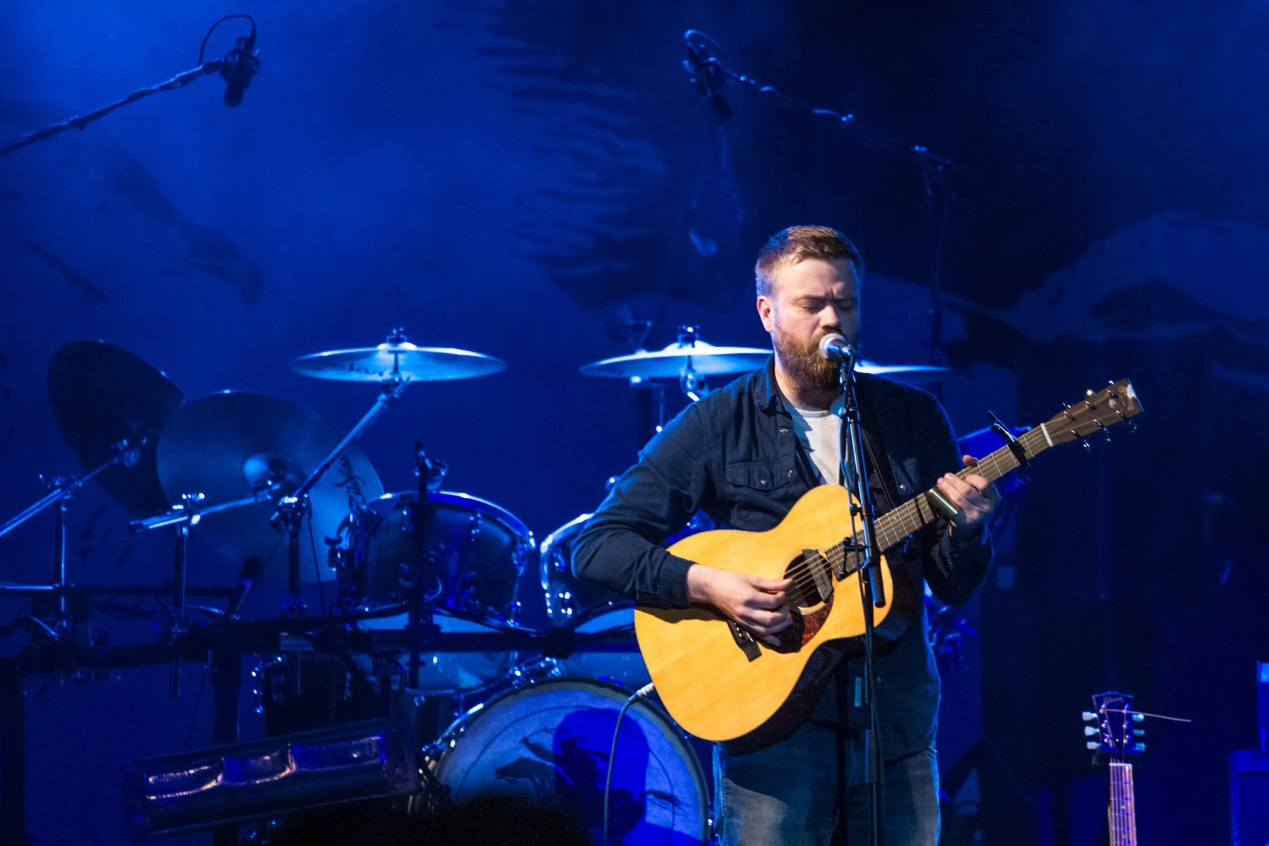 Male musician playing acoustic guitar and singing into a microphone on stage, with drums and skeleton decoration in the background, illuminated by blue stage lighting.