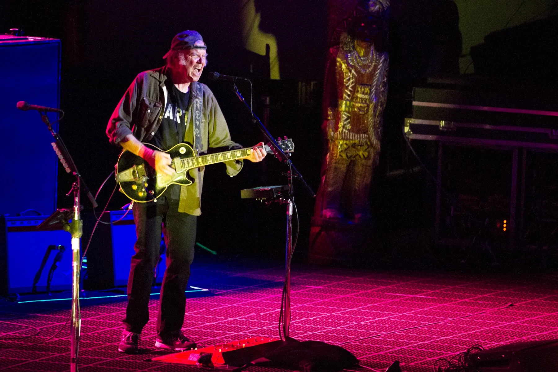 A man passionately sings and plays an electric guitar on stage, surrounded by colorful stage lights and musical equipment.