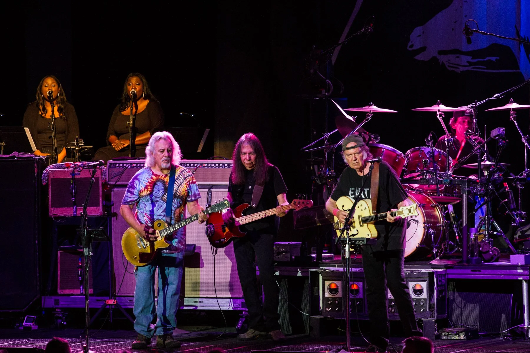 A band performing live on stage, with three guitarists in the foreground and two singers and drummer in the background.