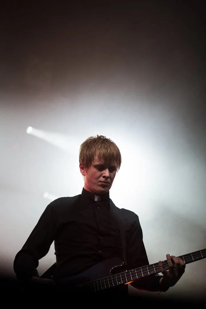 A person dressed in black clothing, possibly a music performer, playing an electric guitar on stage with bright light and smoke effects in the background.