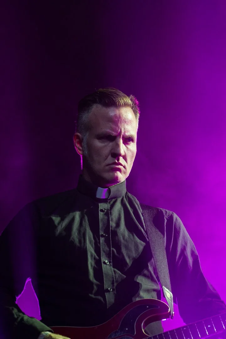 A man dressed as a priest or clergy member wearing a black clerical shirt with a white collar, holding a guitar, against a purple-lit background, with a serious or intense facial expression.