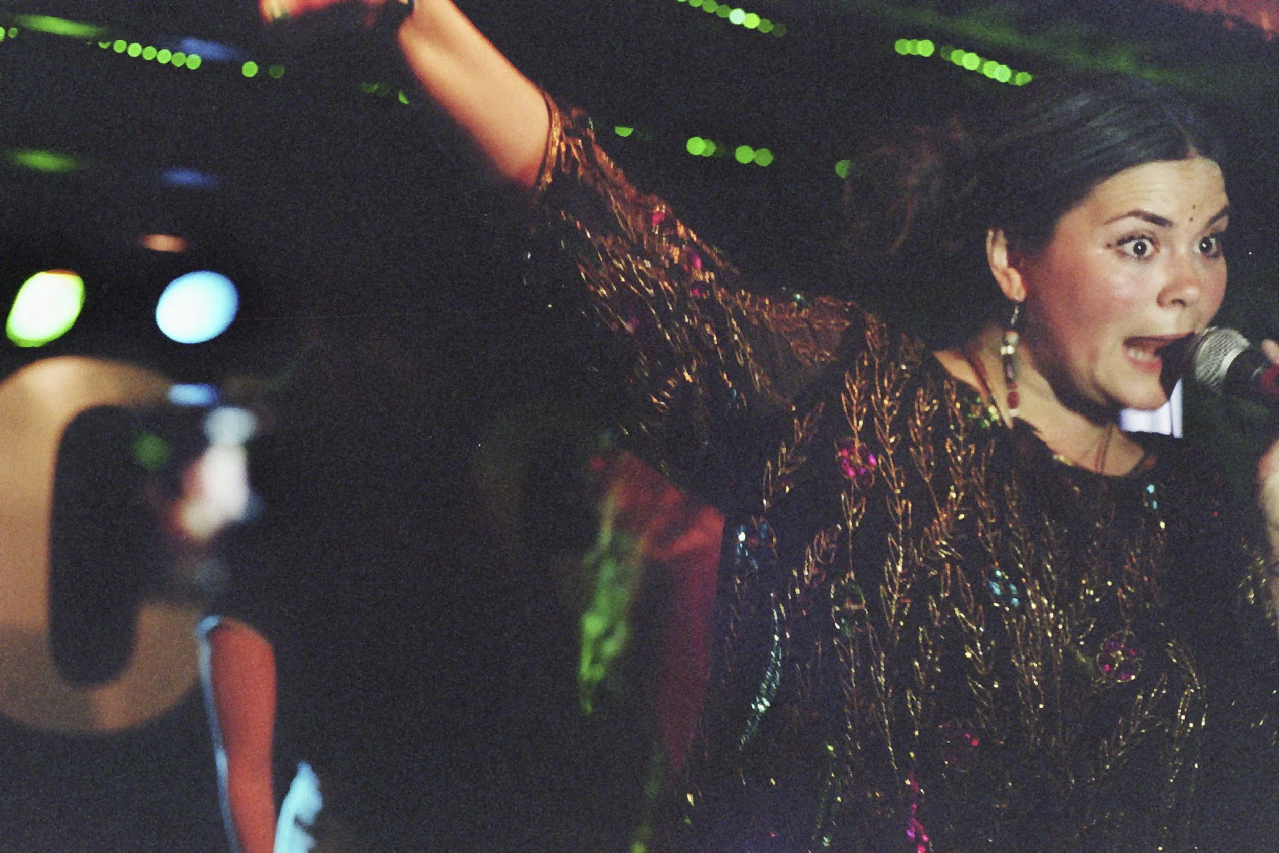 A woman with dark hair singing into a microphone on stage, wearing a dark, colorful, patterned top with lights reflecting off it, and wearing earrings. She has an expressive face and is holding the microphone close to her mouth.