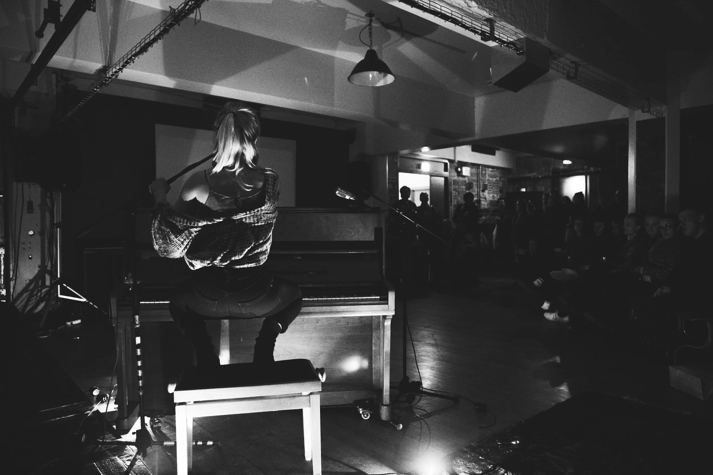 A woman performing on stage with a piano in front of an audience in a dimly lit, cozy venue.