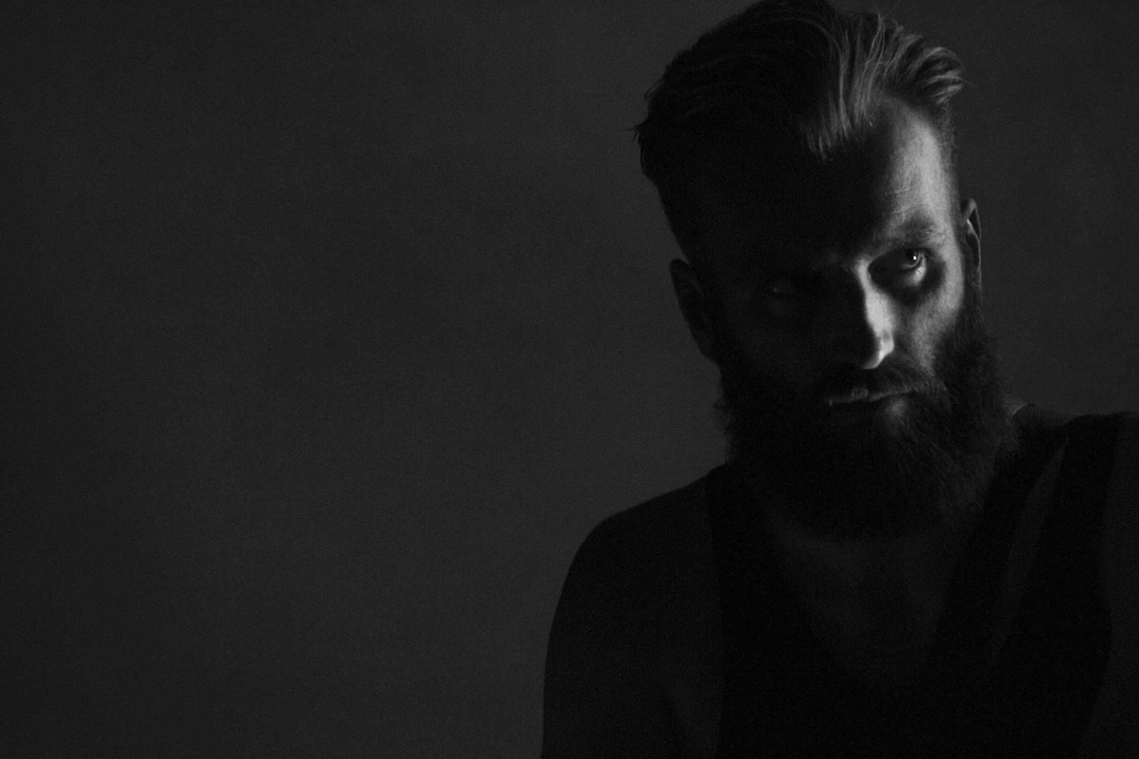 A black and white photo of a man with a beard and styled hair, looking slightly upward, with shadows partially obscuring his face.