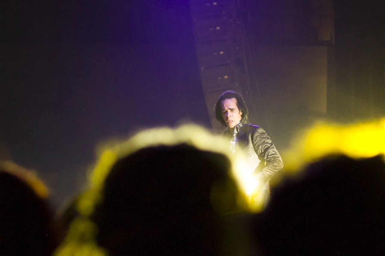 A musician on stage performing with intense expression, illuminated by yellow stage lights, with an audience in the foreground.