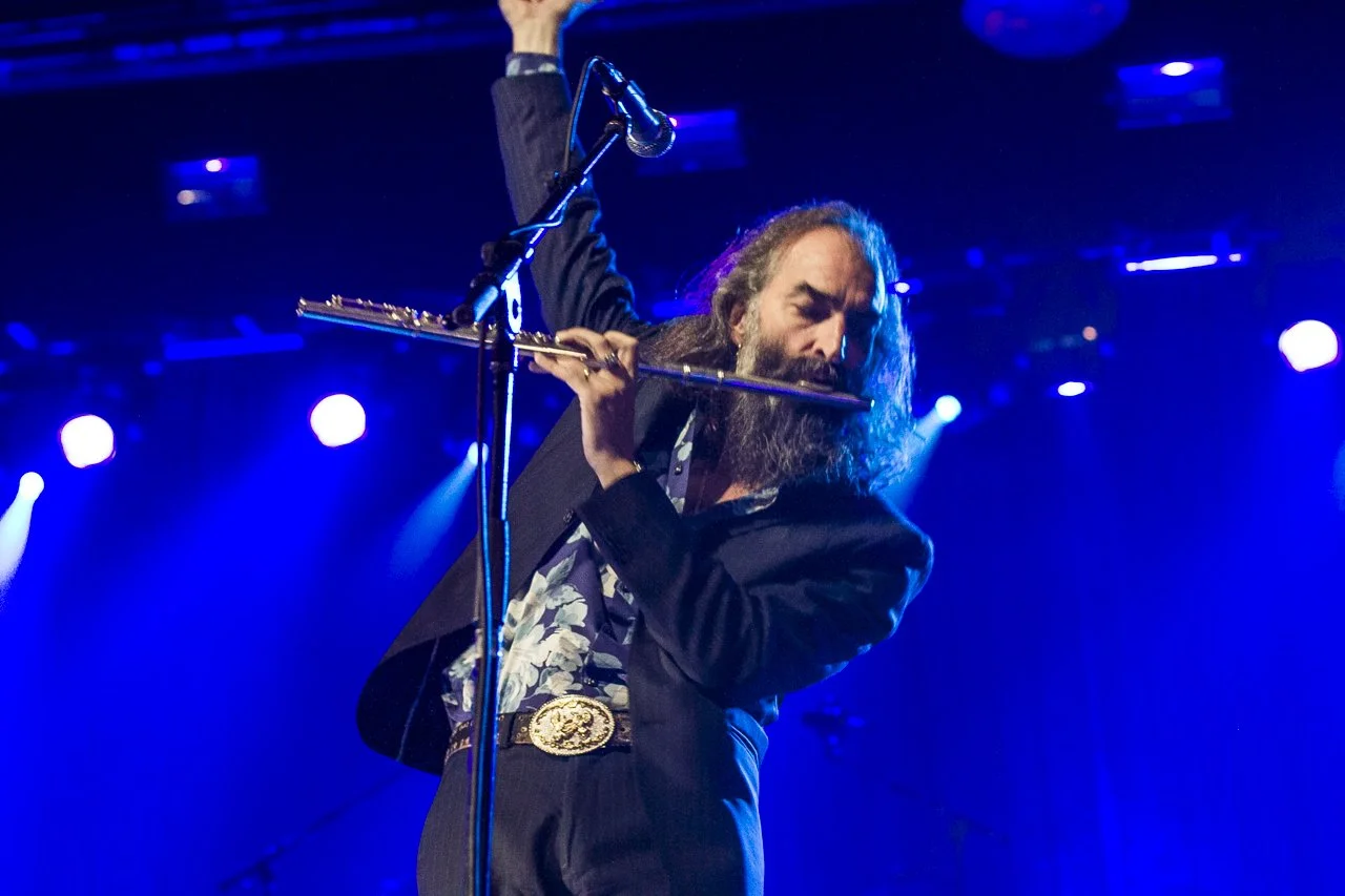A male musician with long hair and a beard playing a flute on stage with blue lighting.