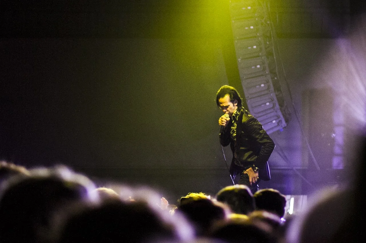 A male performer with long dark hair, wearing a black jacket with gold details, singing into a microphone on stage under yellow and purple lighting, in front of an audience.