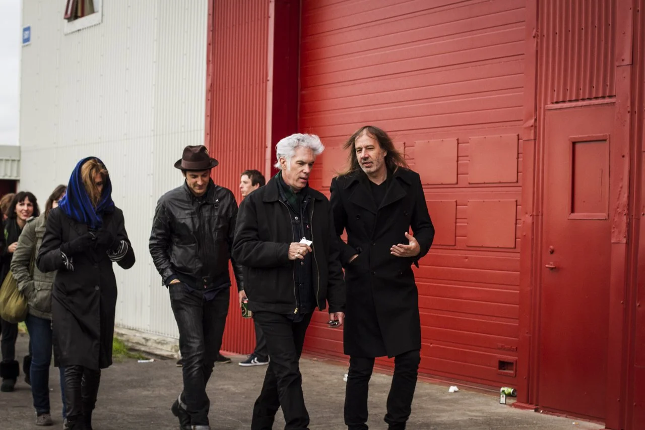 Group of people walking outdoors near a red building, featuring four men in black jackets, and a woman with a dark coat and a headscarf, with some people smiling in the background.