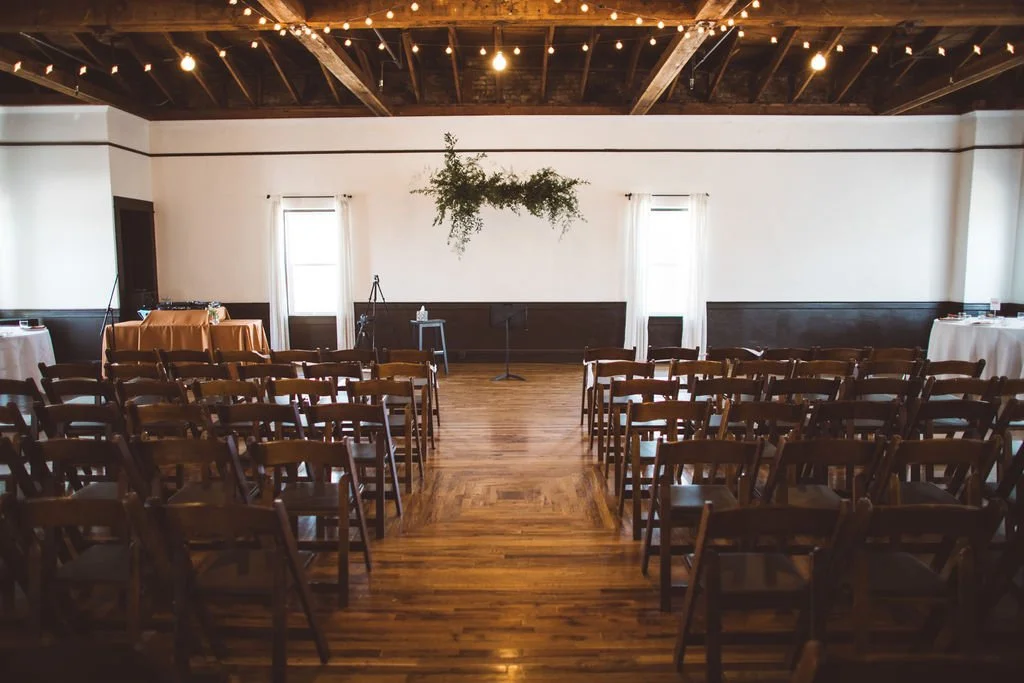 Indoor wedding ceremony in Portland Oregon with ceremony chairs and a ceremony arch. Natural light flows through the windows and exposed rafters and cafe lights give the venue a romantic and intimate charm.