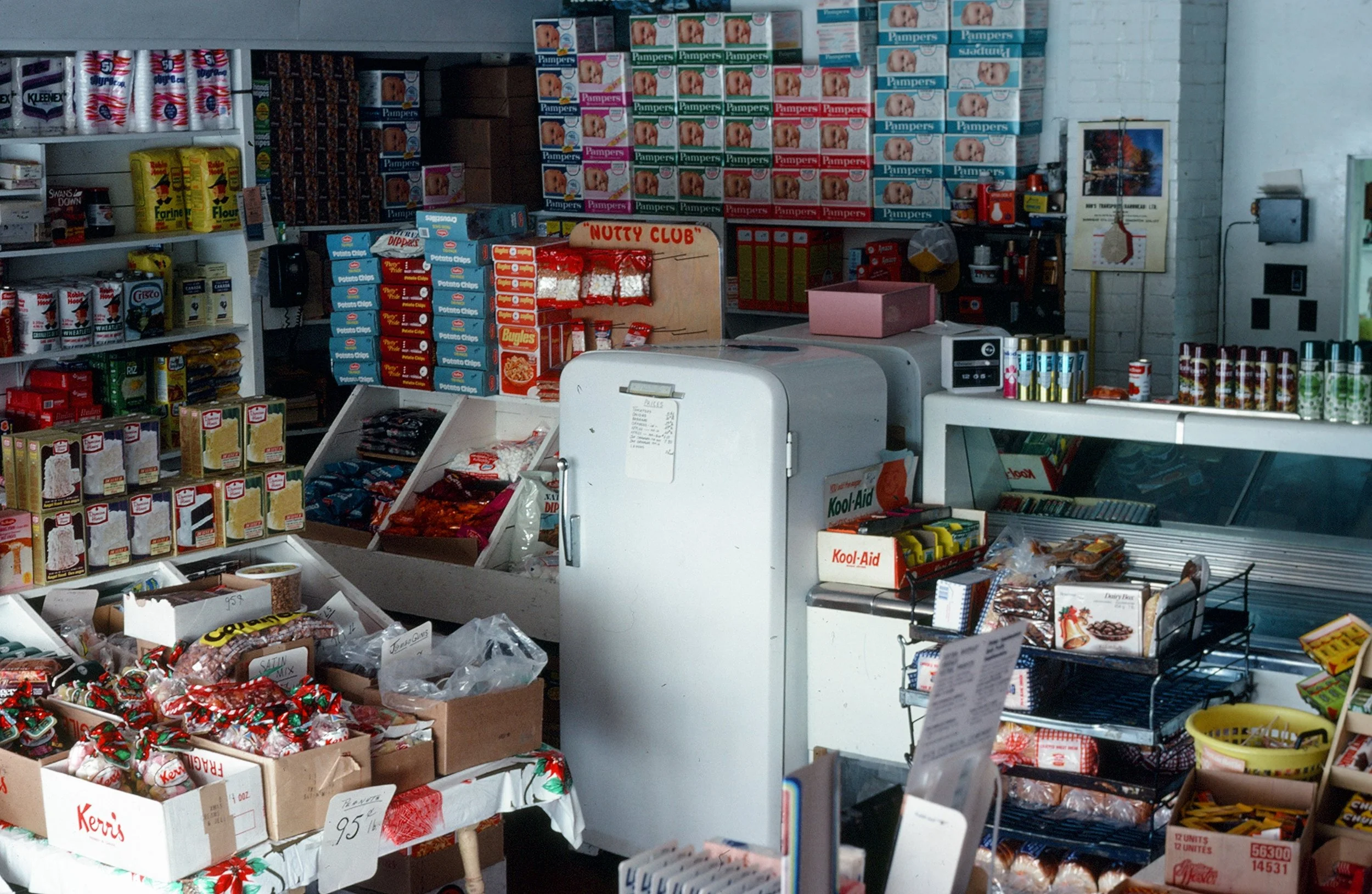 Inside a general store, circa late 1970s, 1980s. Features a white fridge surrounded by products for sale