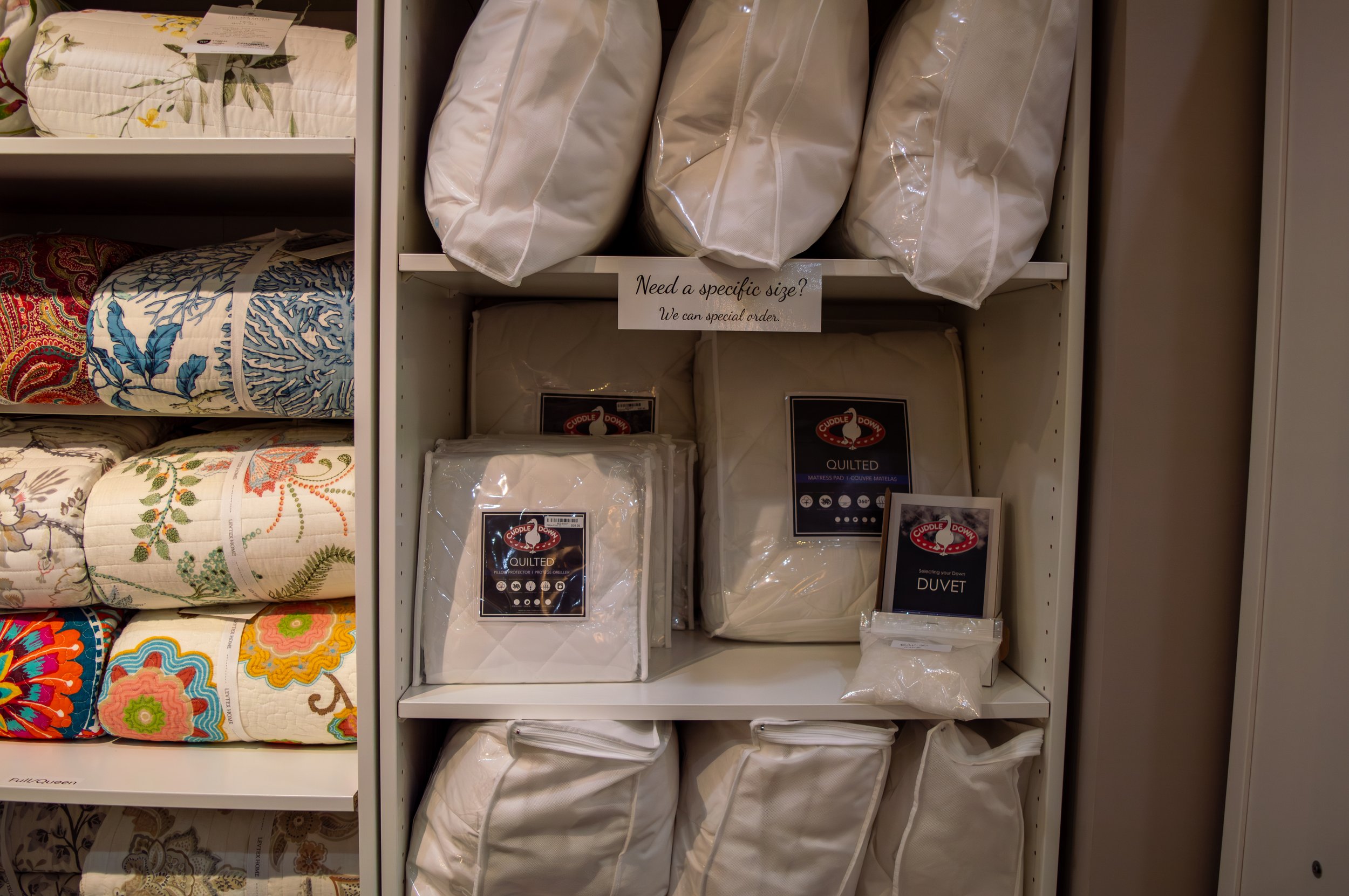 Linens, including packages of down duvets, and quilts, displayed on a white wooden shelf.