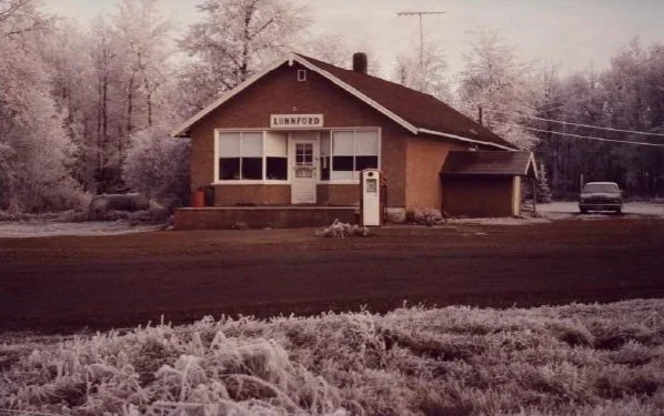 The Lunnford store, with snow covered trees surrounding it, the dirt road in front of the store, and a single gas pump.