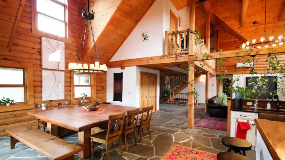 Interior view of a rustic wooden cabin with a large dining table, chandelier, and a loft area. The room features wooden walls and ceiling, stone flooring, and potted plants for decoration.