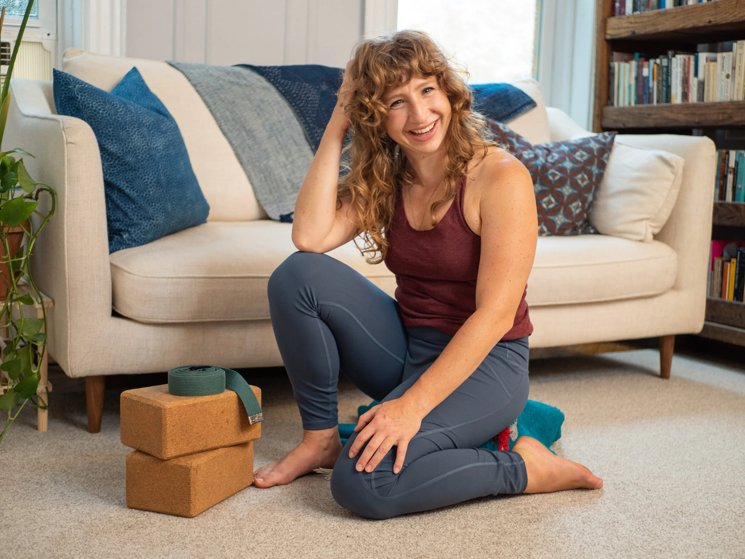 A smiling woman with curly red hair sitting on the floor of a living room, practicing yoga or stretching, with a yoga block and yoga strap nearby.