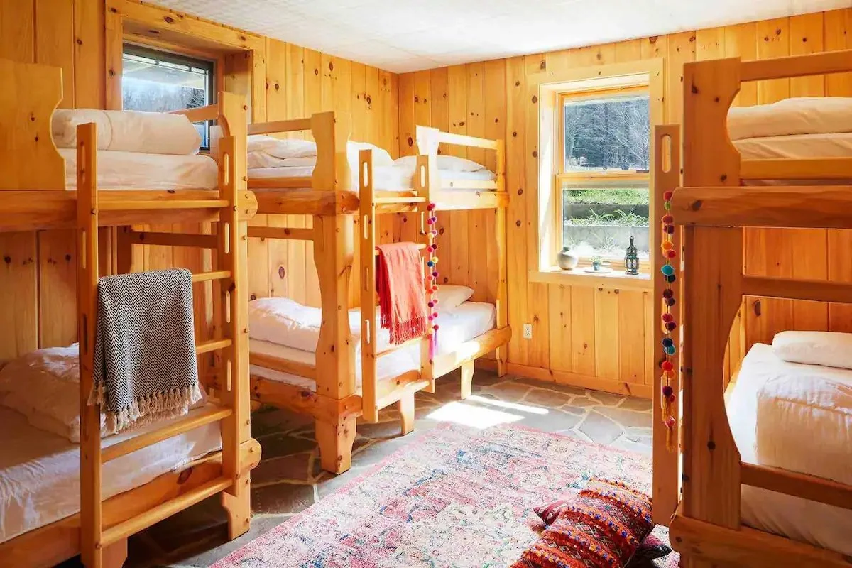A cozy bedroom with wooden walls decorated with bunk beds, each with white bedding and small pillows. The room has two windows allowing natural light to fill the space, with colorful decorations hanging from the beds and a patterned rug on the stone floor.