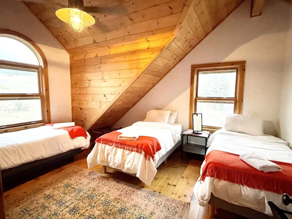 A cozy bedroom with wooden walls and ceiling, three beds with white bedding and red throws, two windows with wooden frames, and a patterned rug on the wooden floor.