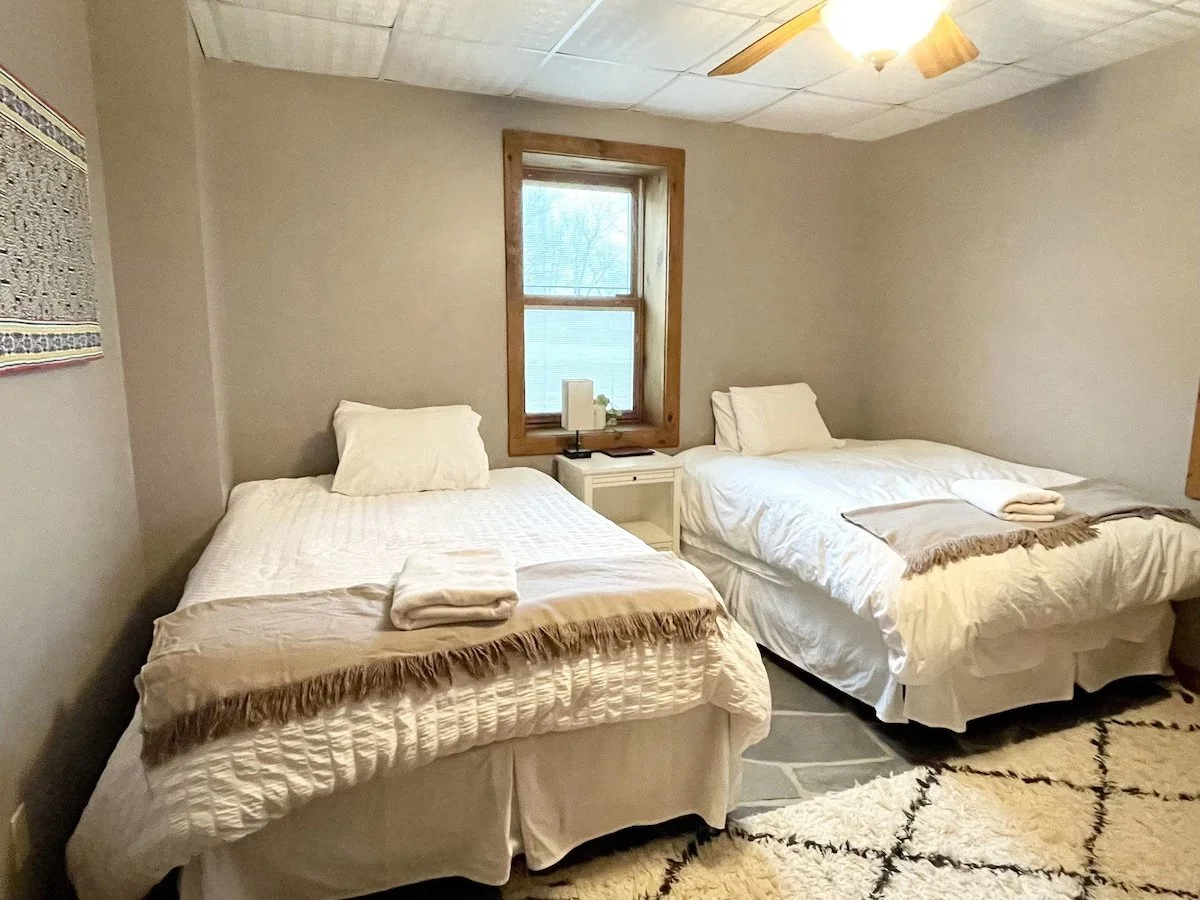 A bedroom with two beds, each with white bedding and a folded towel on top. There is a window between the beds with a small white nightstand beneath it, holding a white lamp and some small items. The ceiling has a fan and light fixture, and a rug is partially visible on the floor.