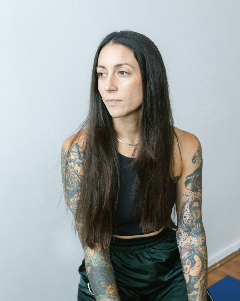 A woman with long dark hair and tattoos on her arms, wearing a black sleeveless top and black pants, standing against a light gray wall, looking to her left.