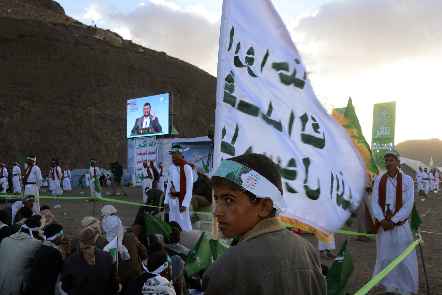  A Yemeni boy raises a flag with a Houthi slogan as the leader, Abdul Malik al Houthi, speaks from a loud screen about their political agenda and the Prophet Mohammad. 
