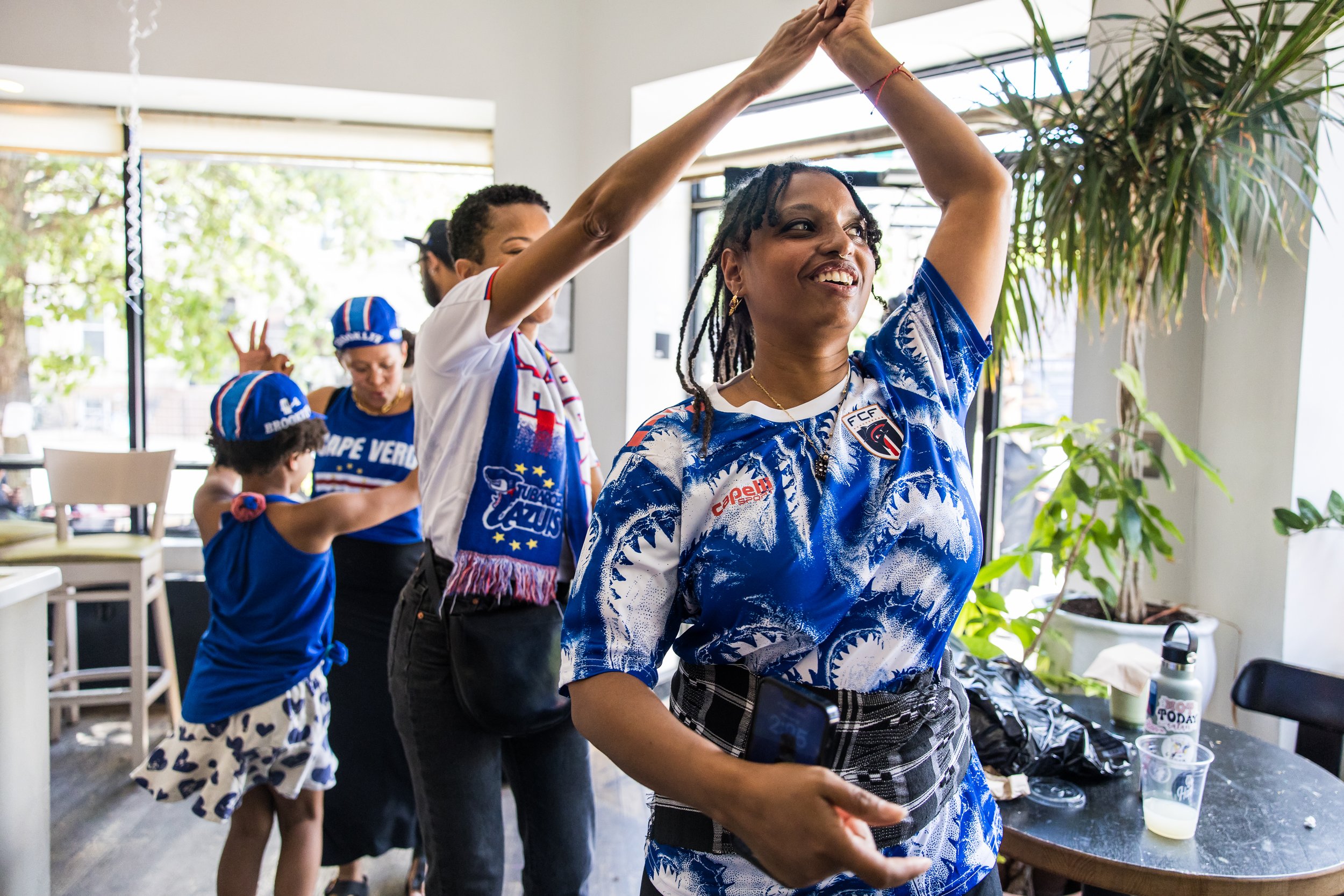  Sara Lopes and Ana Samedo join the celebratory dance party in the cafe. Sara is wearing tchabeta (waist cloths) which are a Cape Verdean cultural tradition worn during the batuku dance.  Bed-Stuy, Brooklyn, June 15, 2026.  