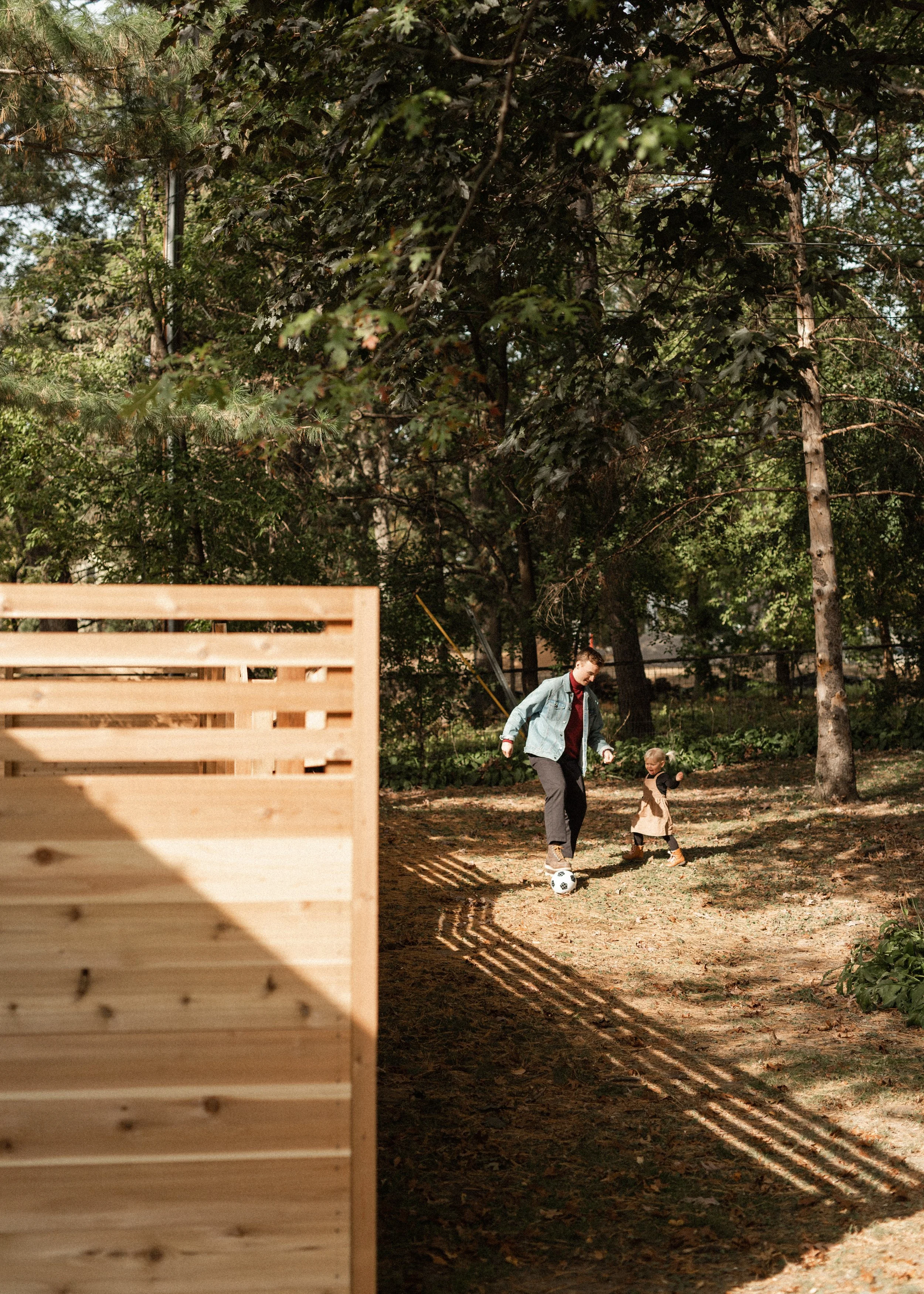 A man and a young girl playing soccer outdoors in a wooded area during daytime, with a wooden fence visible in the foreground.