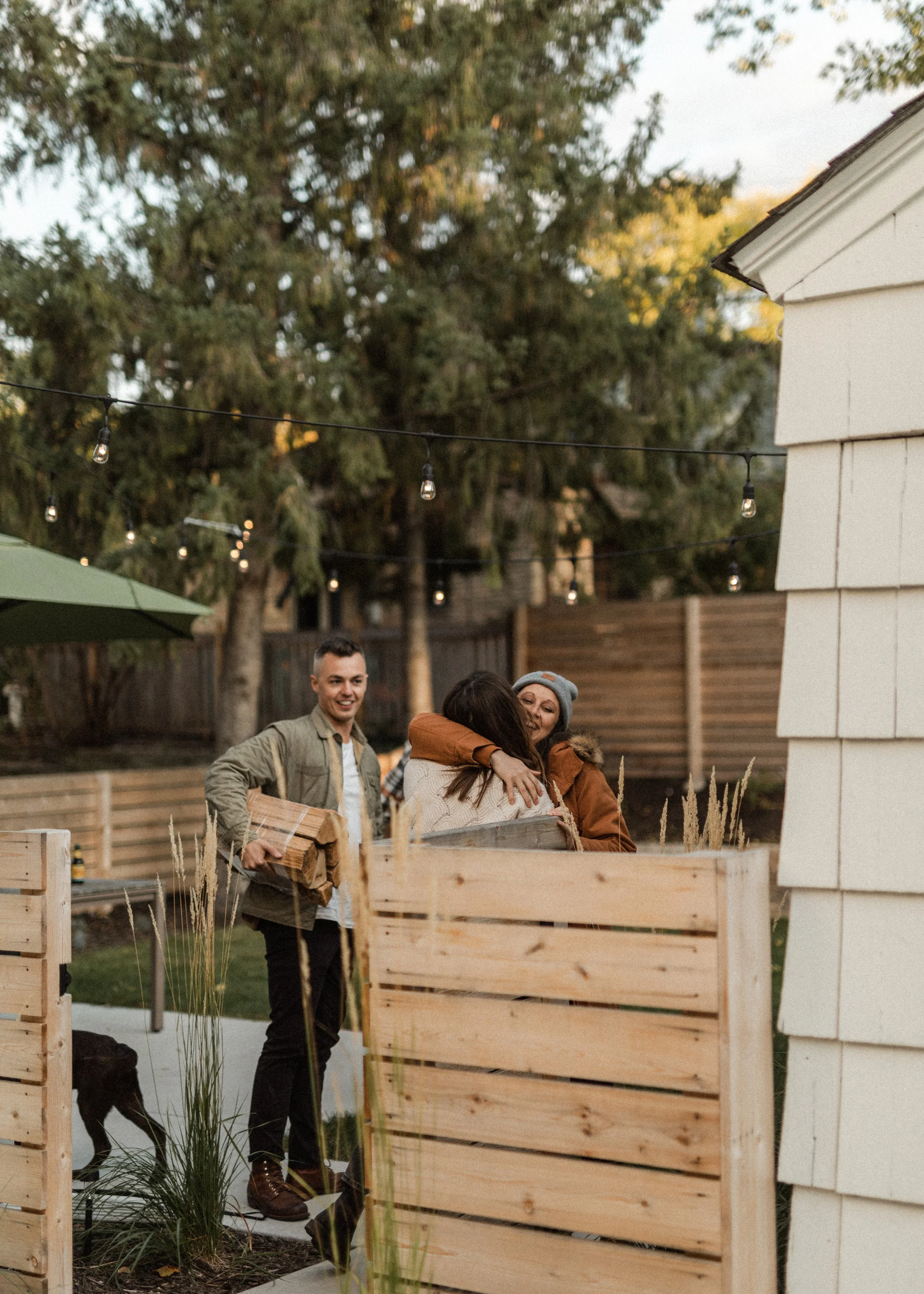 People hugging near a wooden fence in a backyard during dusk, with string lights hanging and trees in the background.
