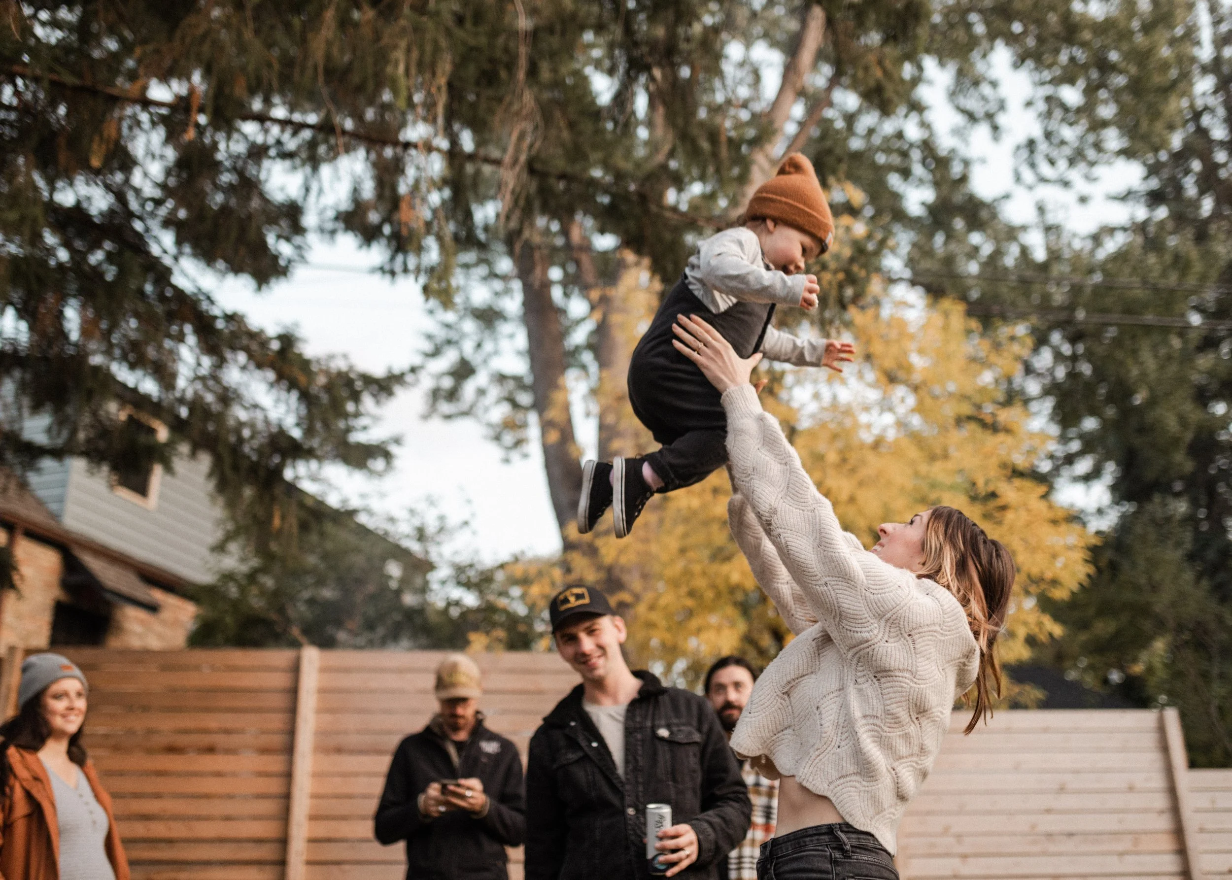 A woman lifts a young boy into the air outdoors during fall, with several onlookers watching and trees with yellow leaves in the background.