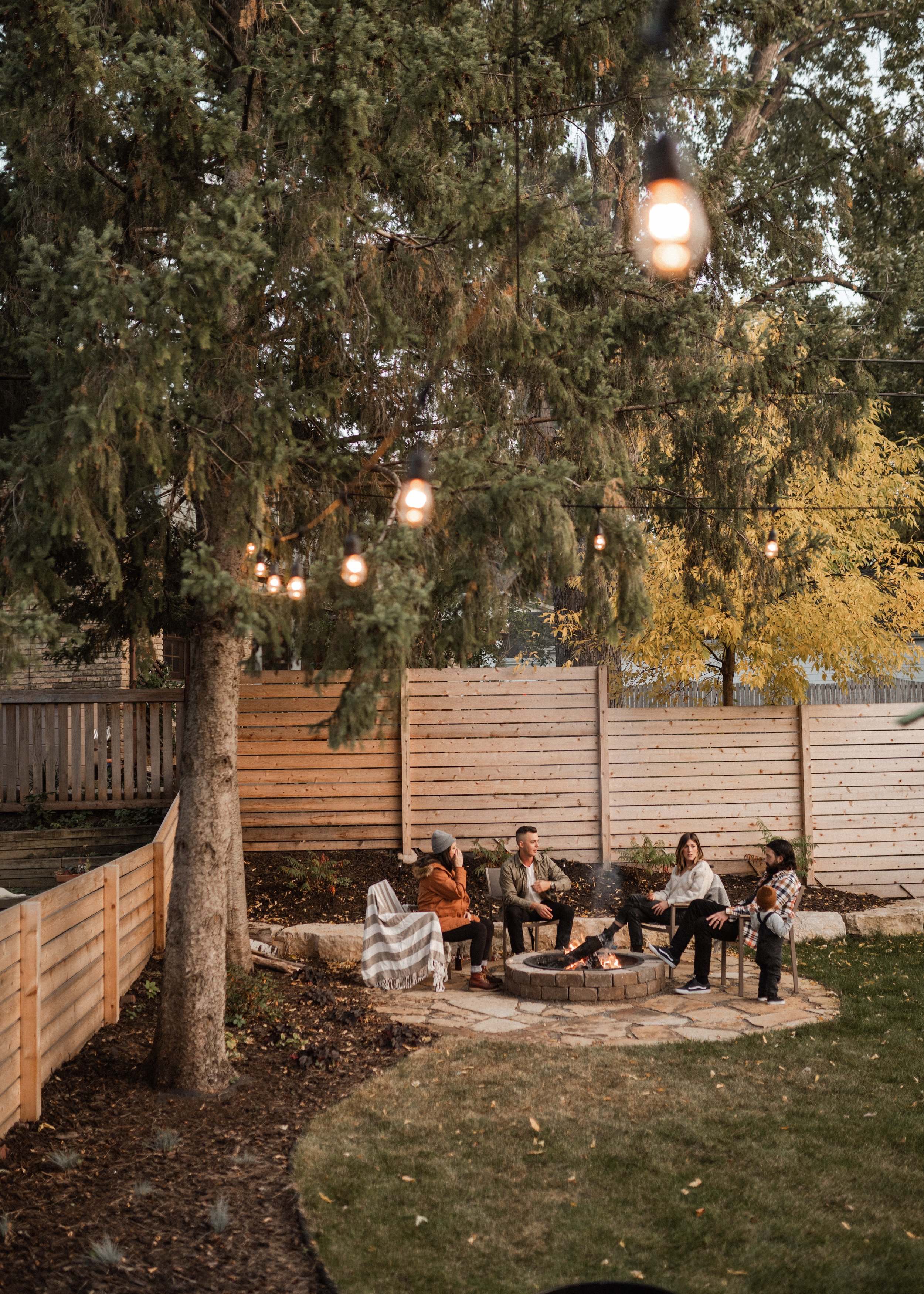 A group of five people gathered around a fire pit in a backyard enclosed by a wooden fence, with string lights hanging from a tree overhead. The scene is set during the evening with warm lighting.