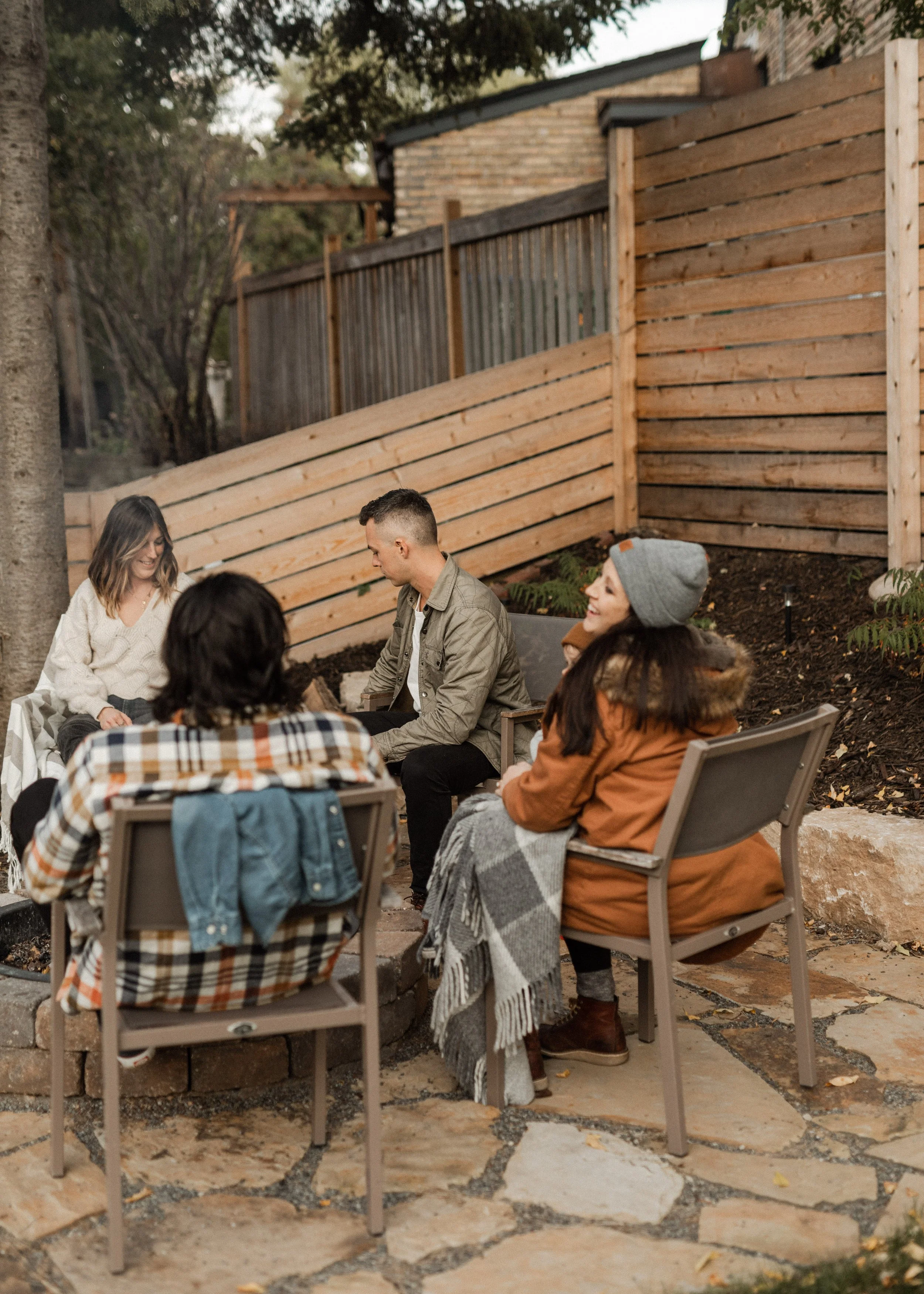 Group of people sitting and talking outdoors in front of a wooden fence, with trees and a brick building in the background, in a garden or backyard setting.