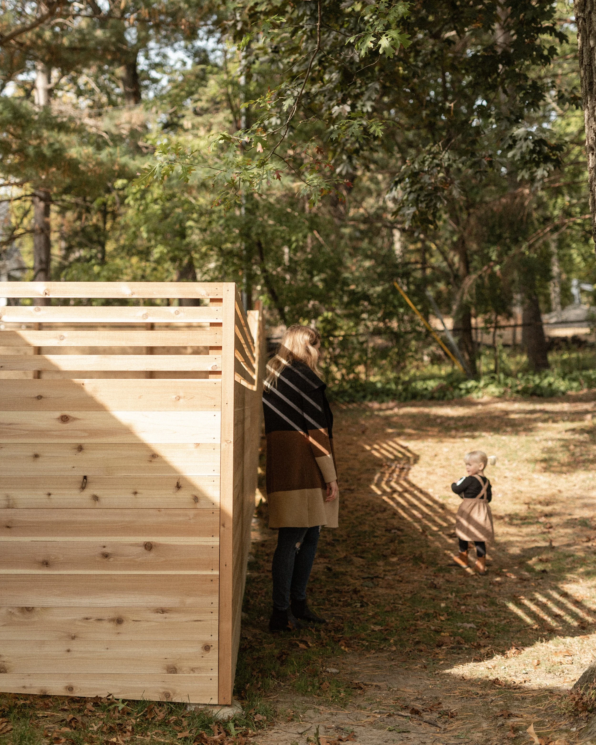 A woman and a child standing outdoors near a wooden fence, with trees and autumn leaves in the background.