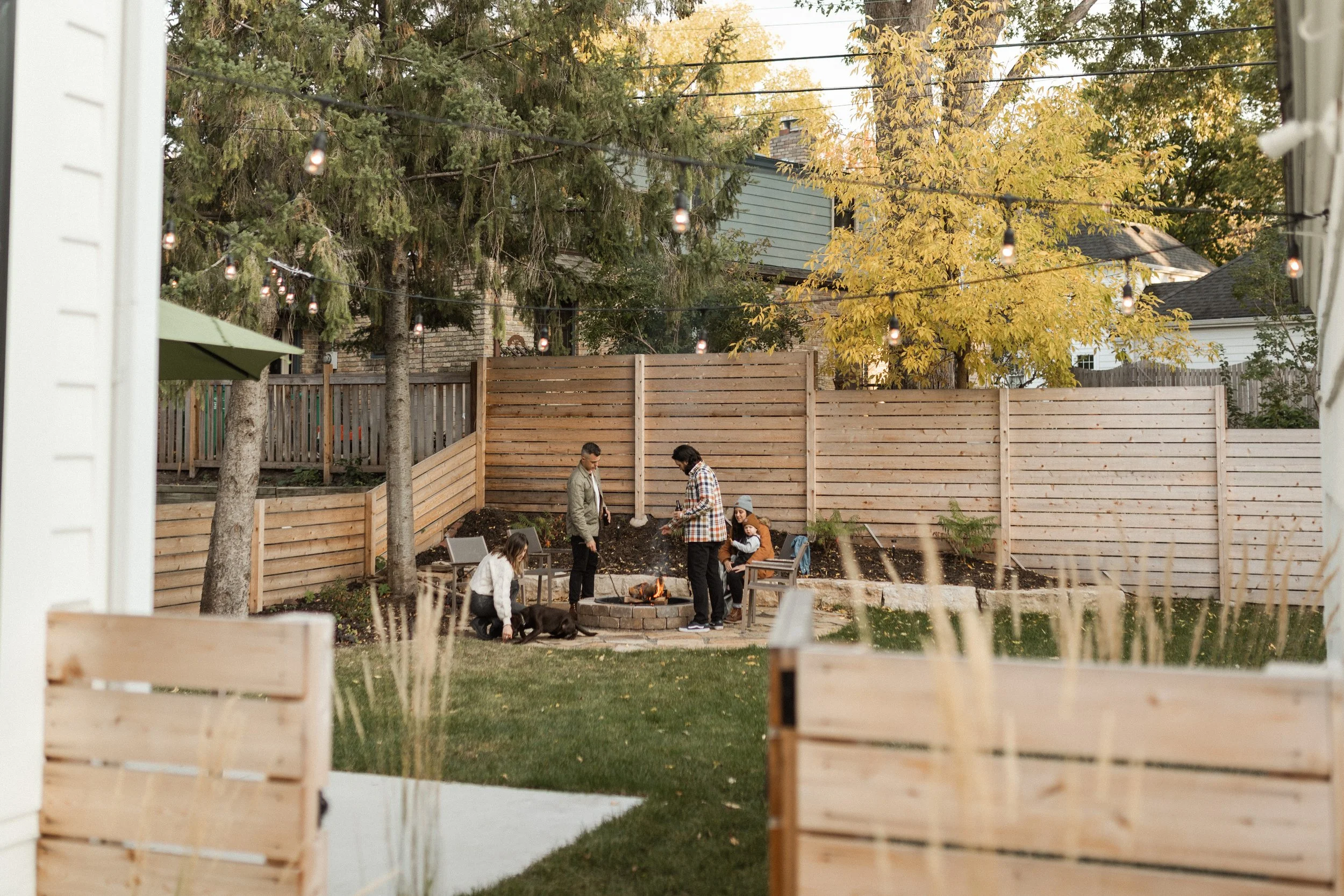 People gathered around a fire pit in a backyard, surrounded by a wooden fence, with trees and houses in the background, decorated with string lights, during autumn.