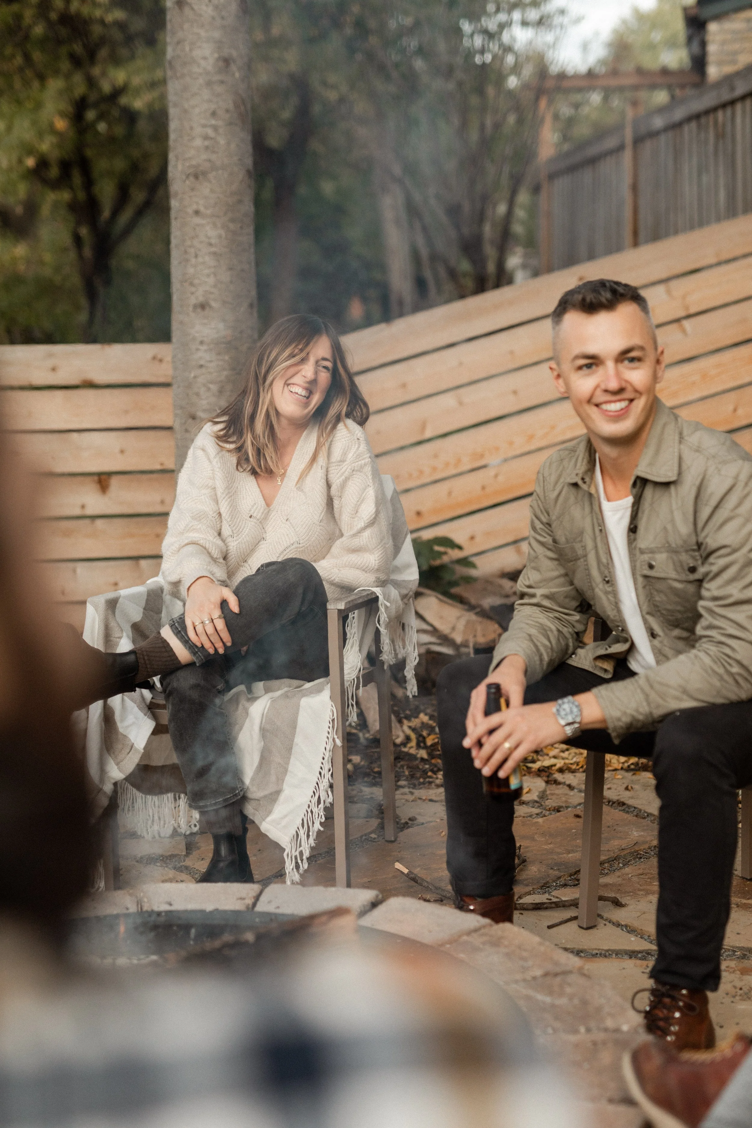 Two smiling people, a woman and a man, sitting outdoors next to a wooden fence and trees. The woman is wearing a cream sweater and has shoulder-length brown hair. The man is wearing a light-colored jacket and is holding a bottle. They are sitting on 