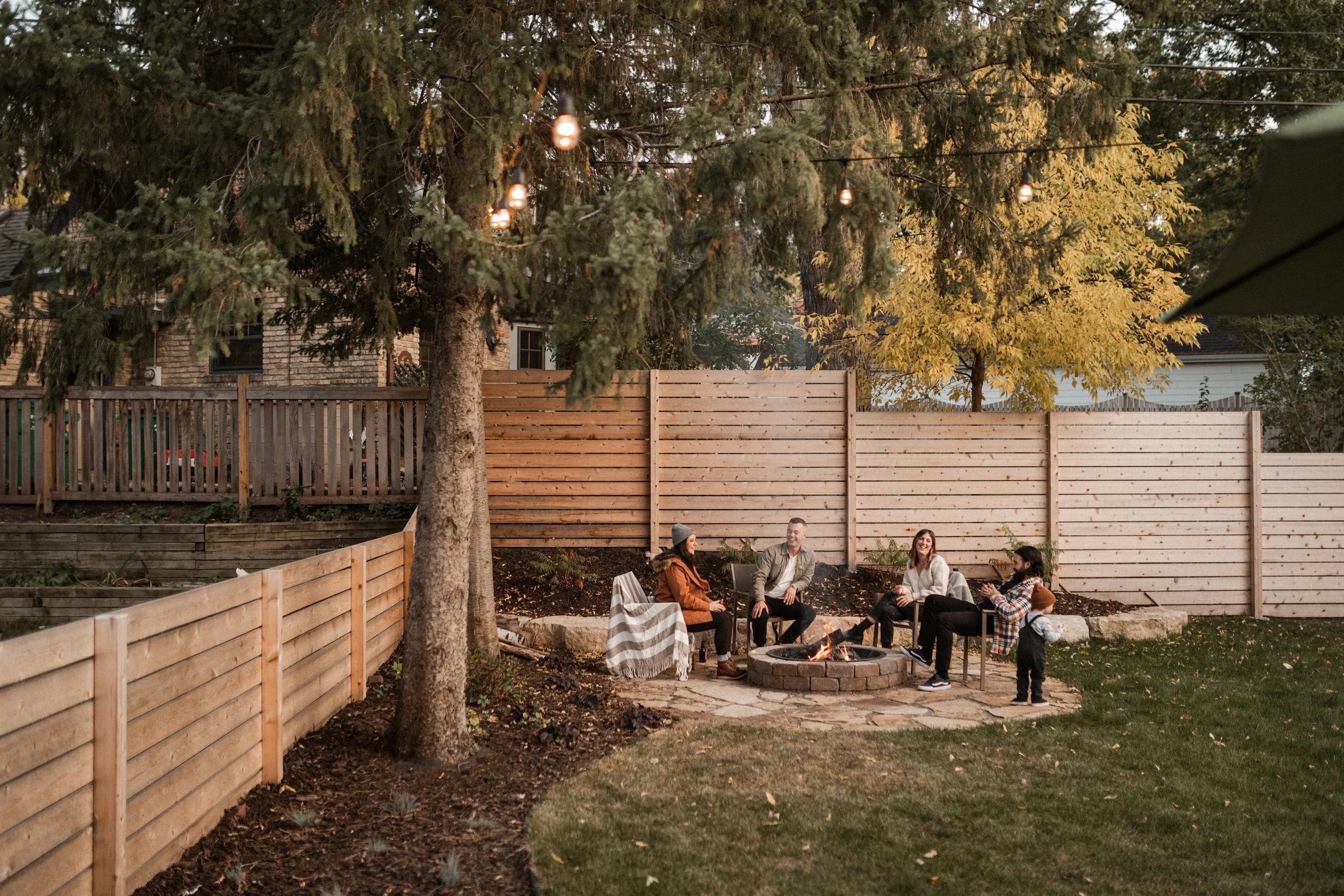 People sitting around a fire pit in a backyard with wooden fencing and trees, some string lights hanging from the trees.