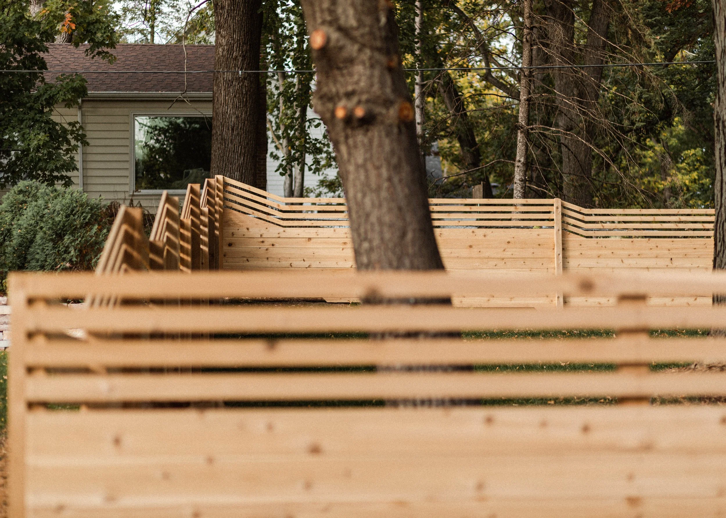 A wooden fence in the foreground with horizontal slats, a large tree trunk in the middle, and a house with a window and a shingled roof surrounded by trees in the background.