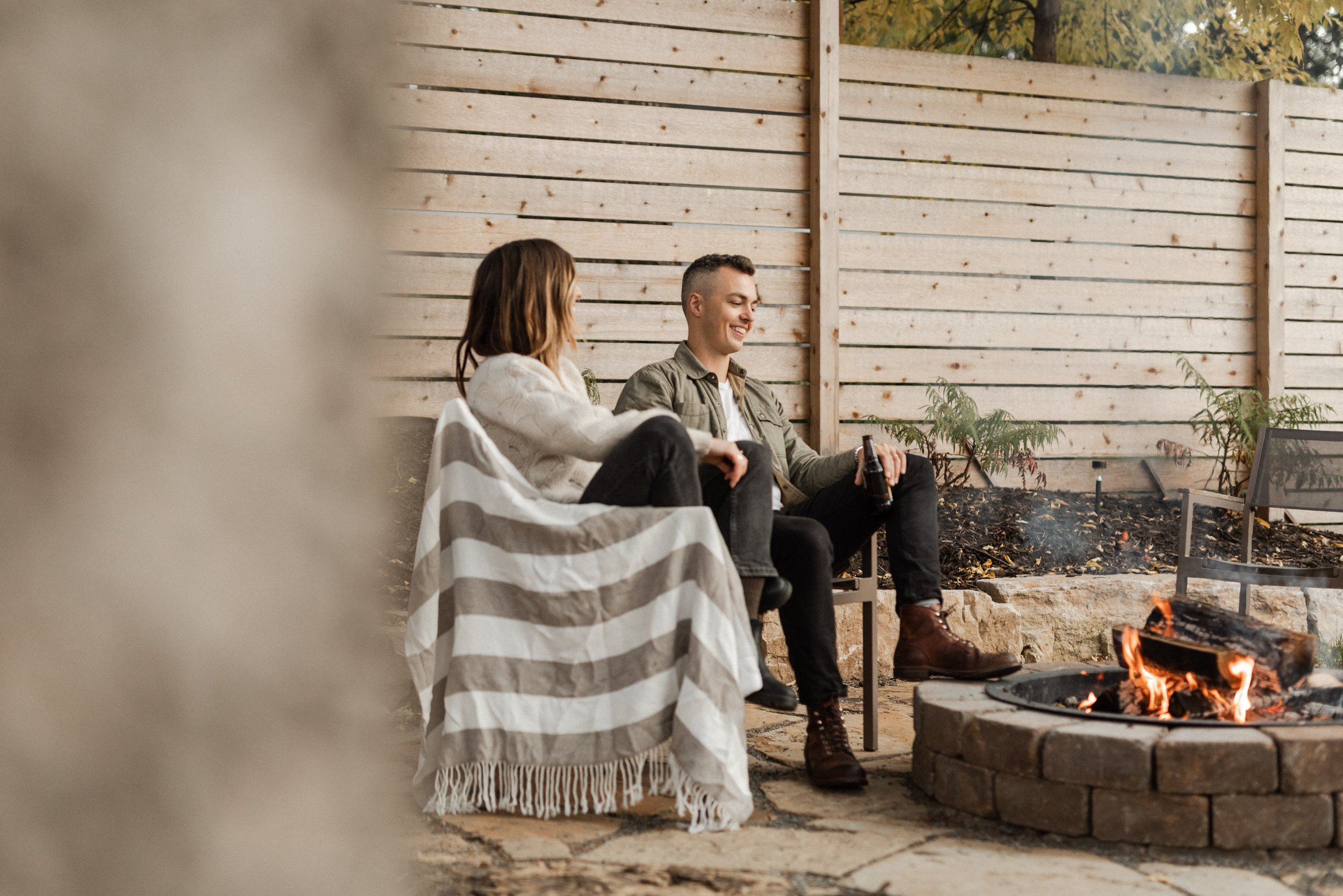 Two people sit by a fire pit in a backyard with a wooden fence. The woman has brown hair and is wearing a white sweater, the man has short hair and is wearing a green jacket. They are smiling and enjoying drinks.