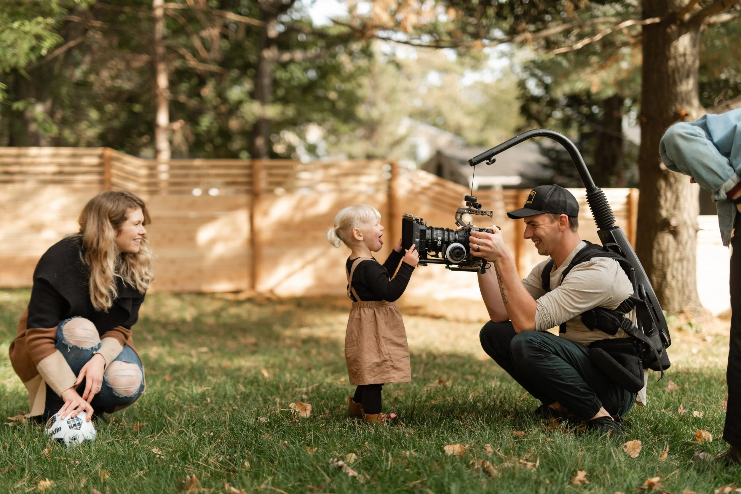A young girl holding a camera with a man kneeling in front of her, smiling and looking at her while a woman kneeling beside them and a person on the right side of the image are watching. They are outdoors in a backyard with trees and a wooden fence i