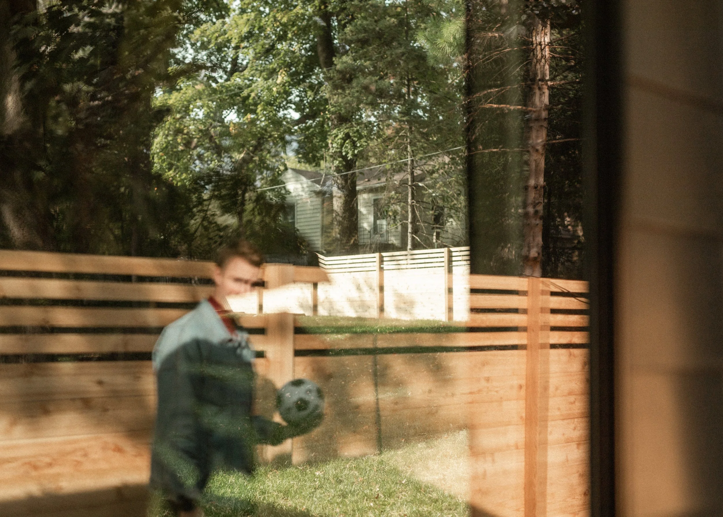 A young boy in a green jacket, standing behind a wooden fence, playing soccer outdoors with a black and white soccer ball. There are trees and a house in the background, and the photo is taken through a window.