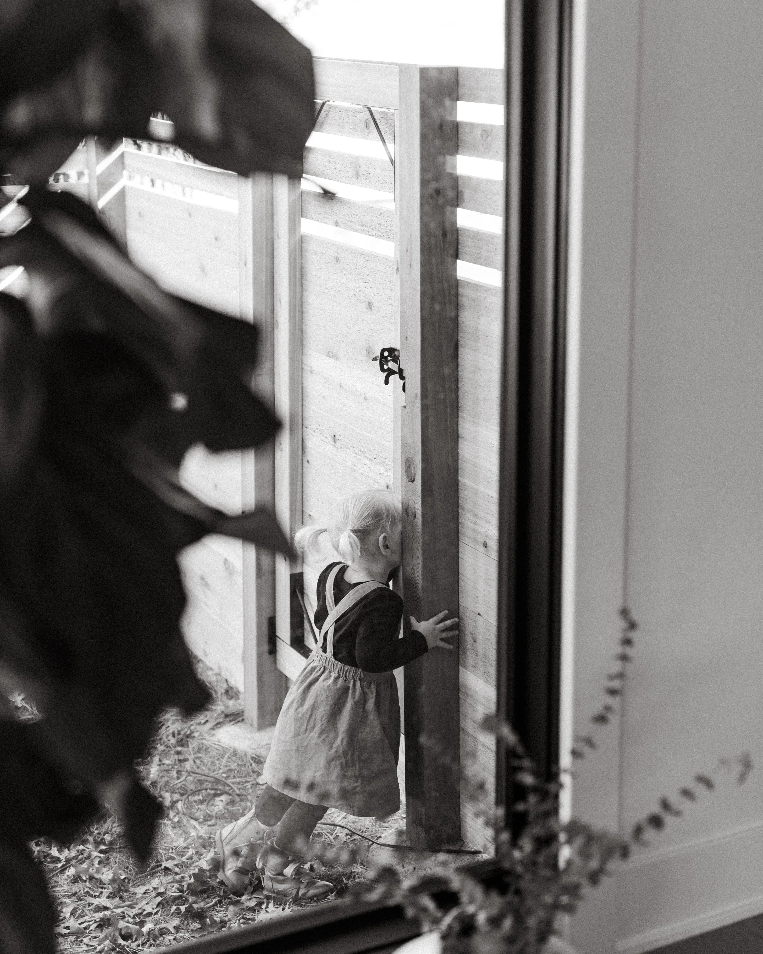 A young girl wearing a dress and sneakers is crouched down peering into a wooden enclosure, possibly at a pet or an animal inside. The image is in black and white.