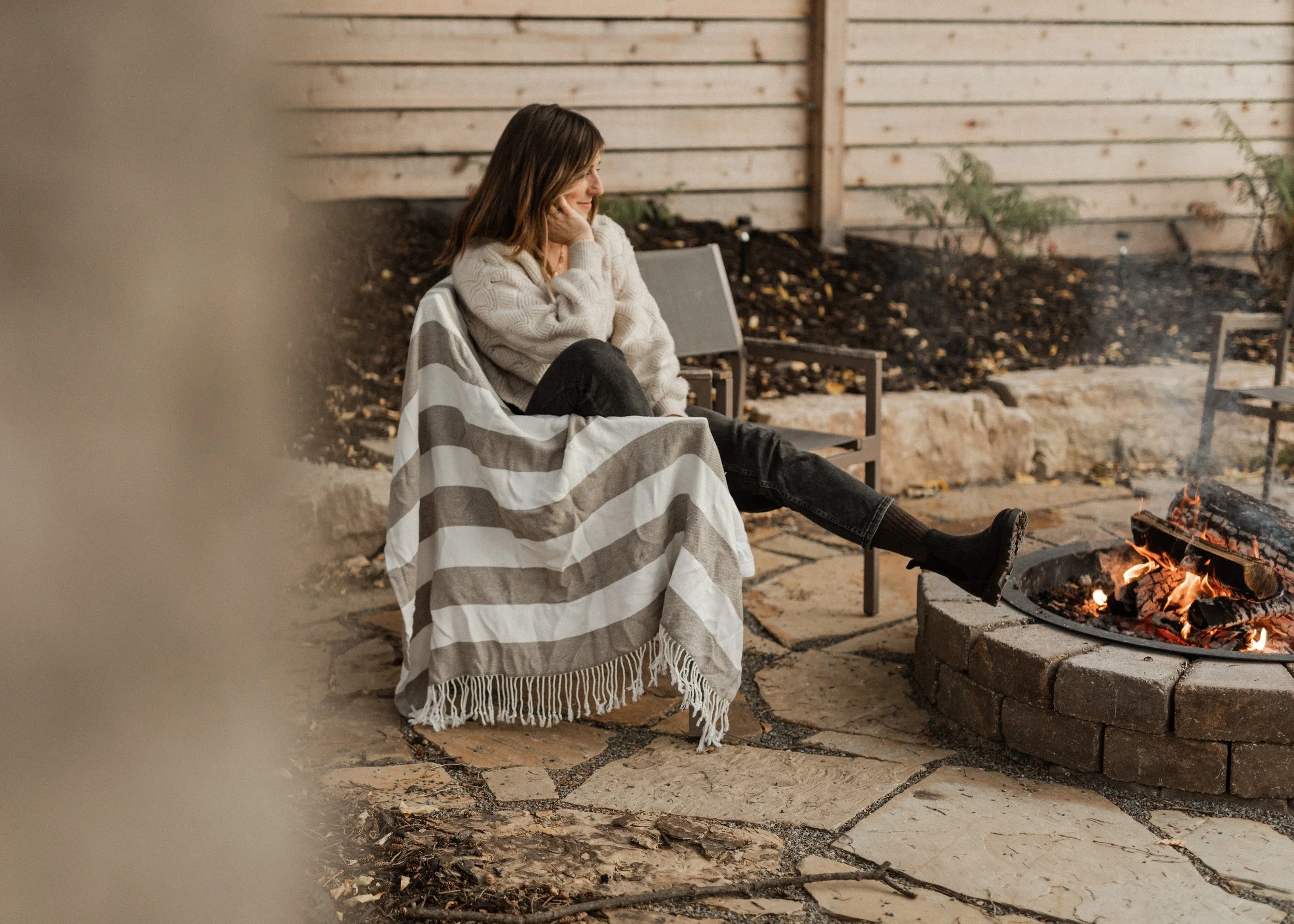 A woman sitting on a bench outdoors near a campfire, wrapped in a striped blanket, looking at the fire, with a wooden fence and some plants in the background.