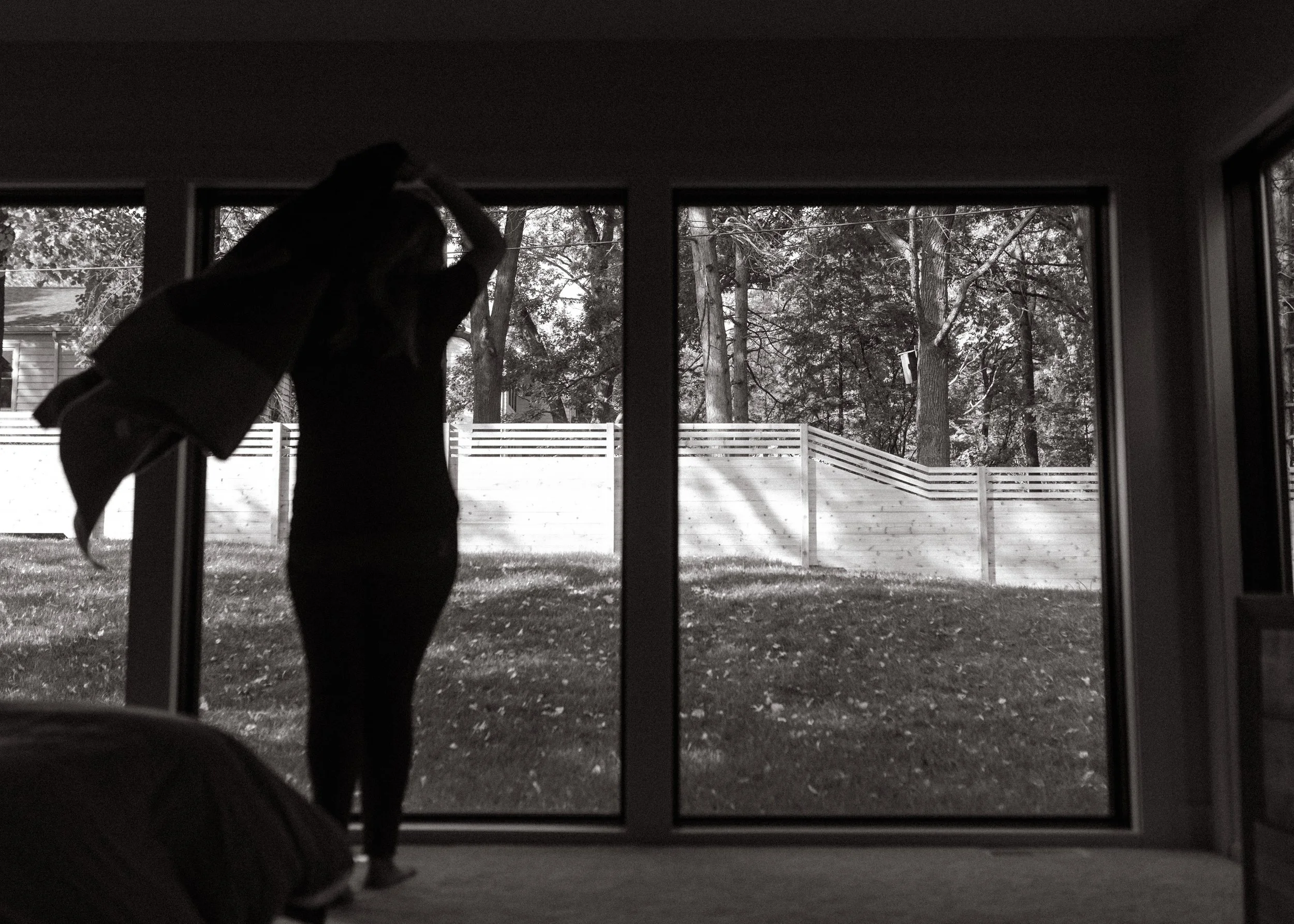 Person standing by large glass doors, looking outside at trees and a fenced yard.