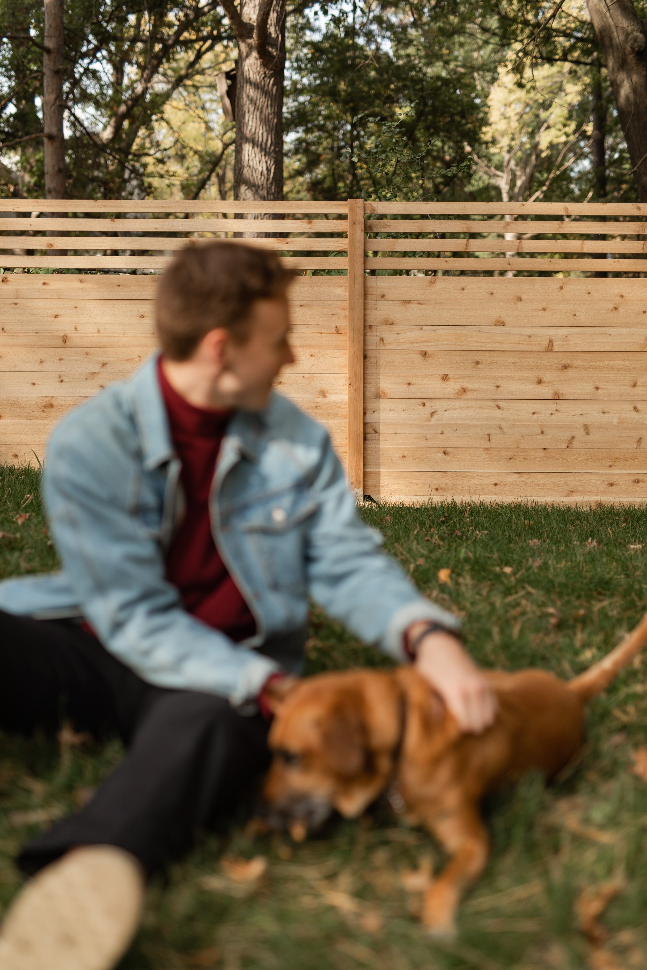 A young man with curly brown hair, wearing a light blue denim jacket, red shirt, and black pants, sitting on grass in front of a wooden fence with trees in the background. He is petting a brown dog with floppy ears.