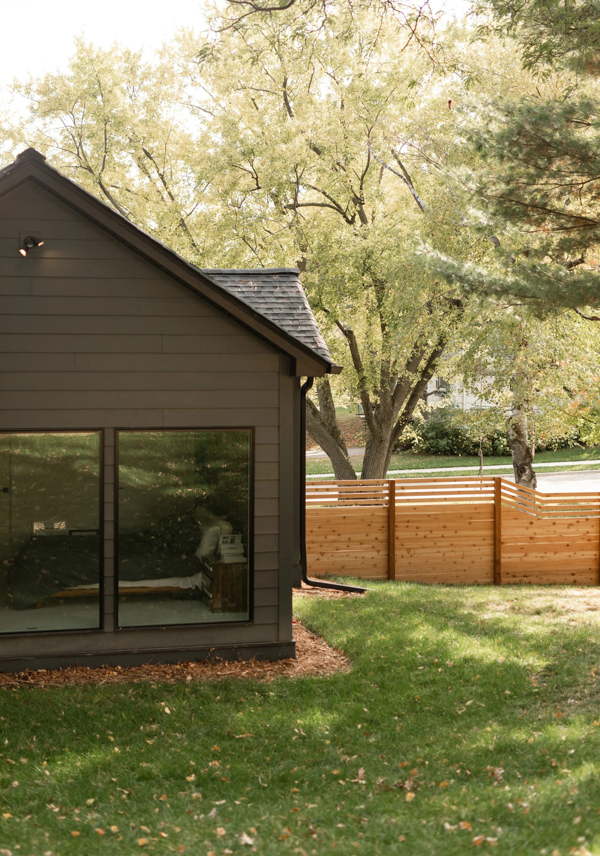 A view of a house with large glass windows, a gray exterior, and a sloped roof, surrounded by green grass and trees with yellow leaves, a wooden fence, and partial view of a street.
