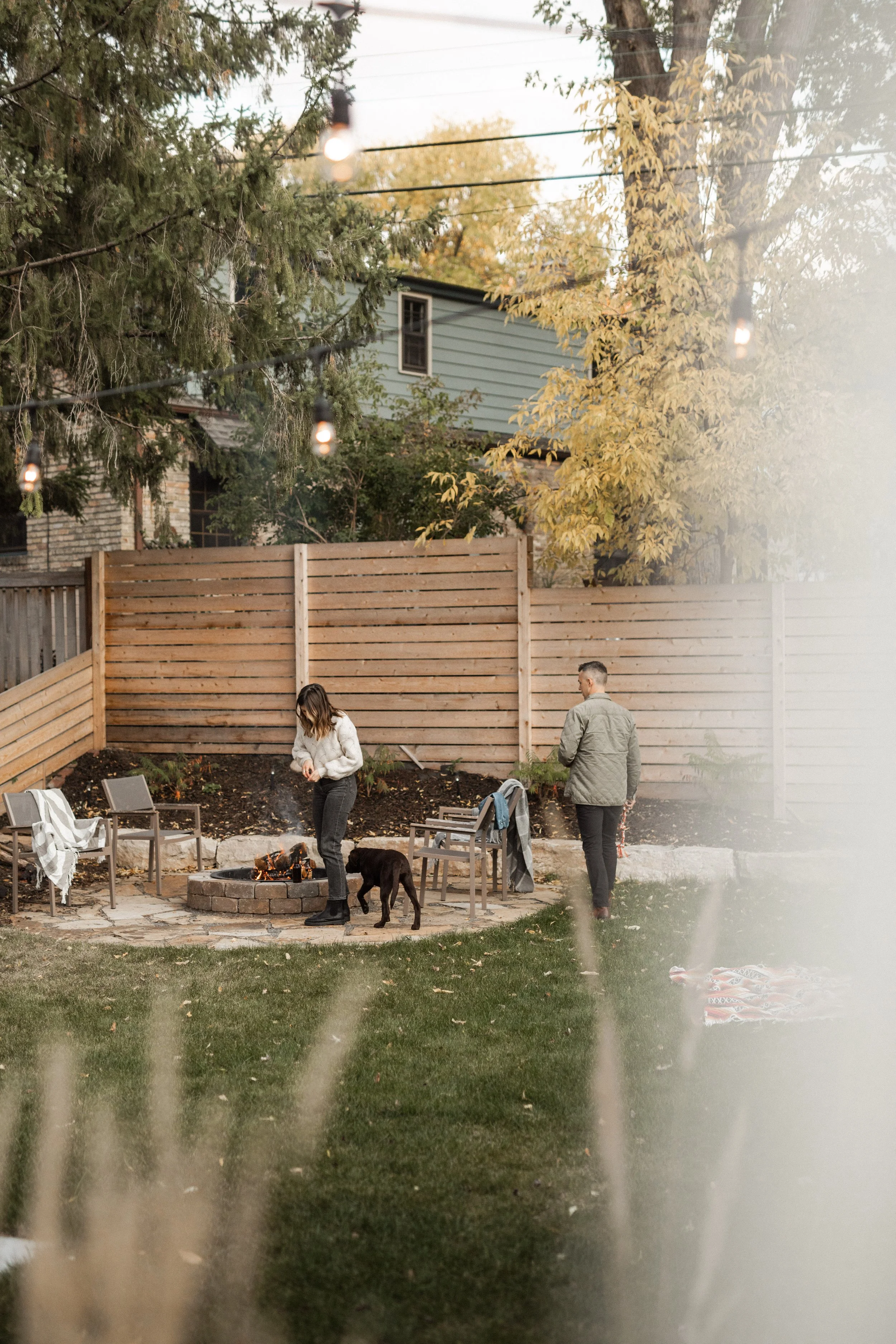 People gathered around a backyard fire pit, a woman with a dog, and a man standing beside her, with a wooden fence, trees, and a house in the background.